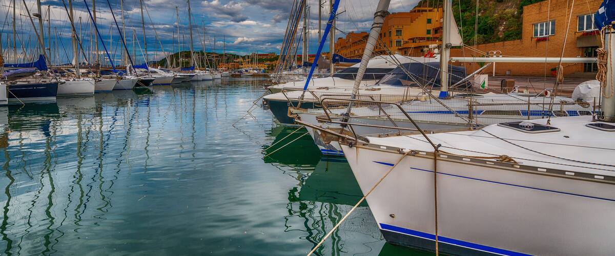A marina with multiple sailboats docked, a building with red and yellow facades, and a calm water body reflecting the sky.