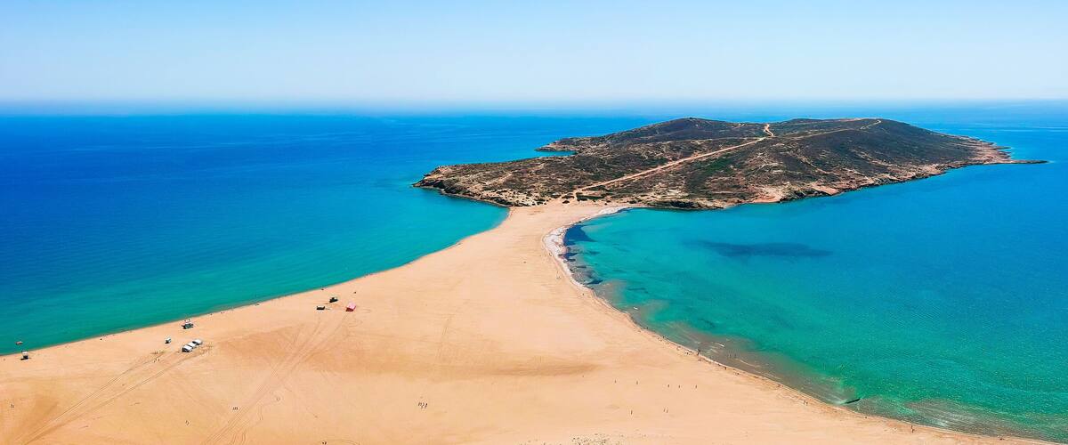 A coastal landscape with a sandy beach, clear turquoise waters, and a small island with a road leading to it.