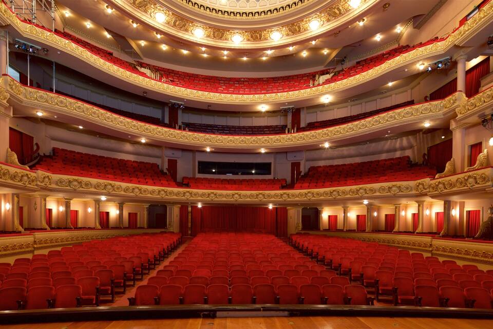 A grand theater with ornate golden railings, red seats, and multiple balconies.