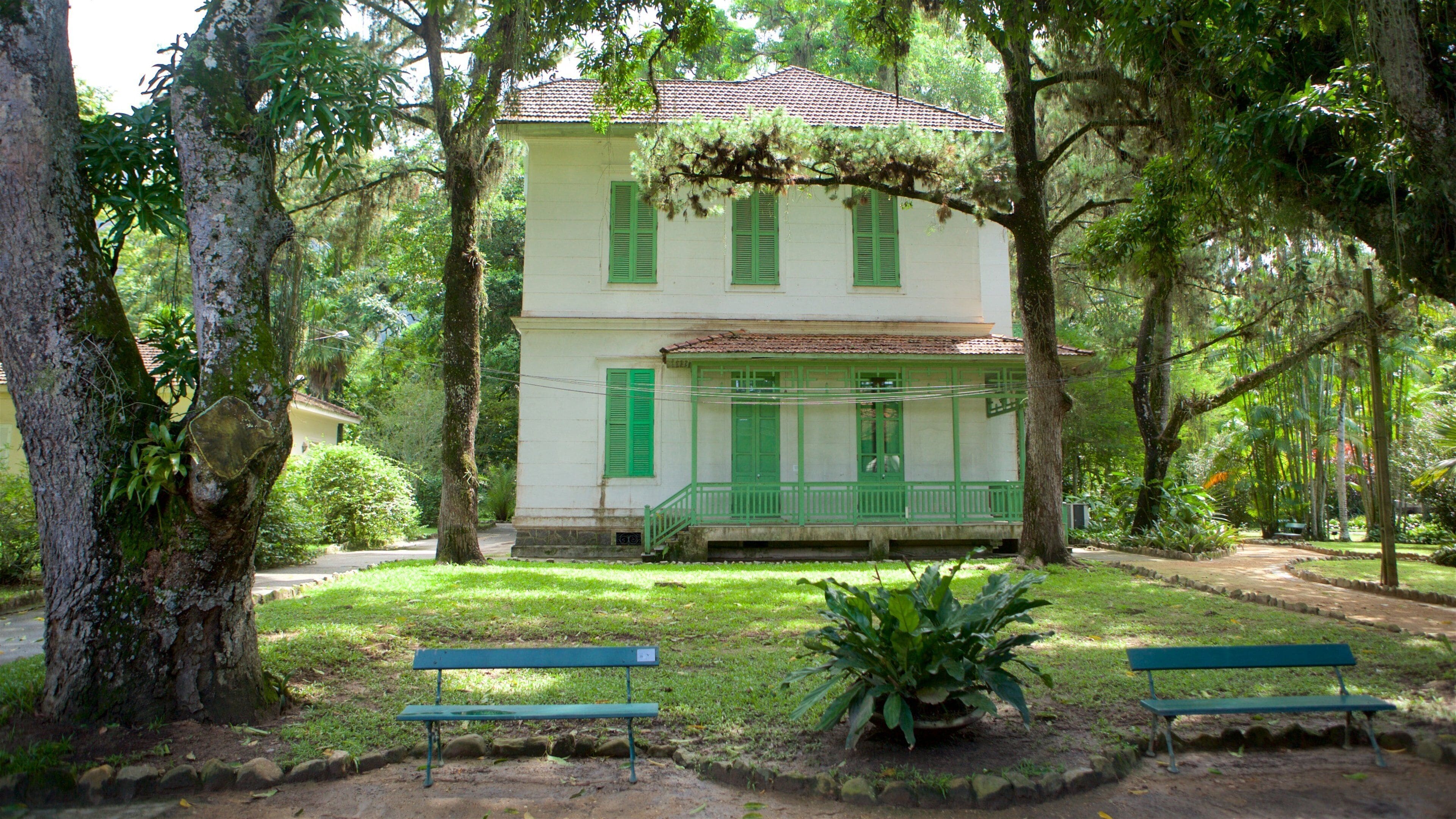 Un bâtiment de style colonial à deux étages, doté de volets verts et d’un porche, entouré d’une végétation luxuriante et d’arbres.