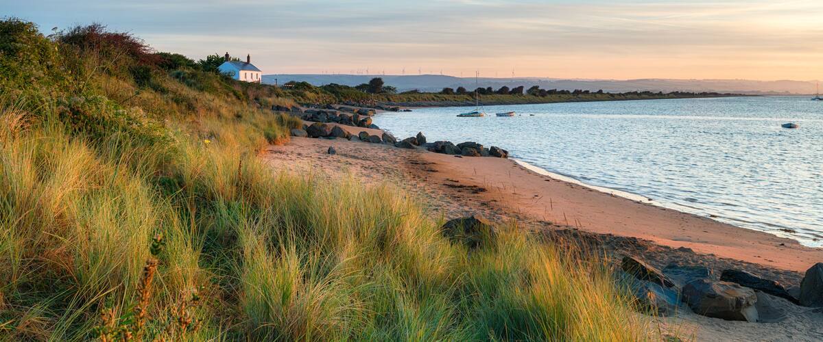 A coastal landscape with a sandy beach, grassy dunes, a small white house, and calm water with boats.