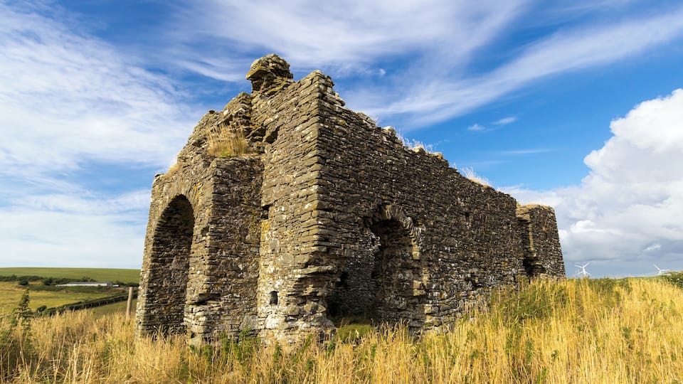 An ancient stone ruin with arched doorways, set against a backdrop of a clear blue sky and wind turbines in the distance.
