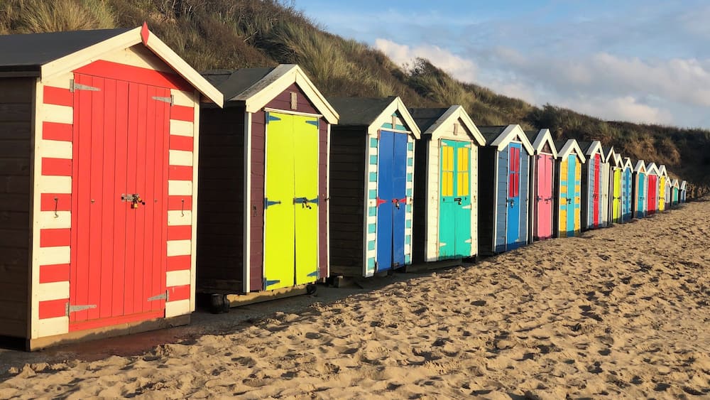 A row of colorful beach huts along a sandy beach with a grassy hill in the background.