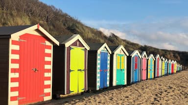Fileira de cabanas coloridas ao longo de uma praia arenosa, com uma colina gramada ao fundo.