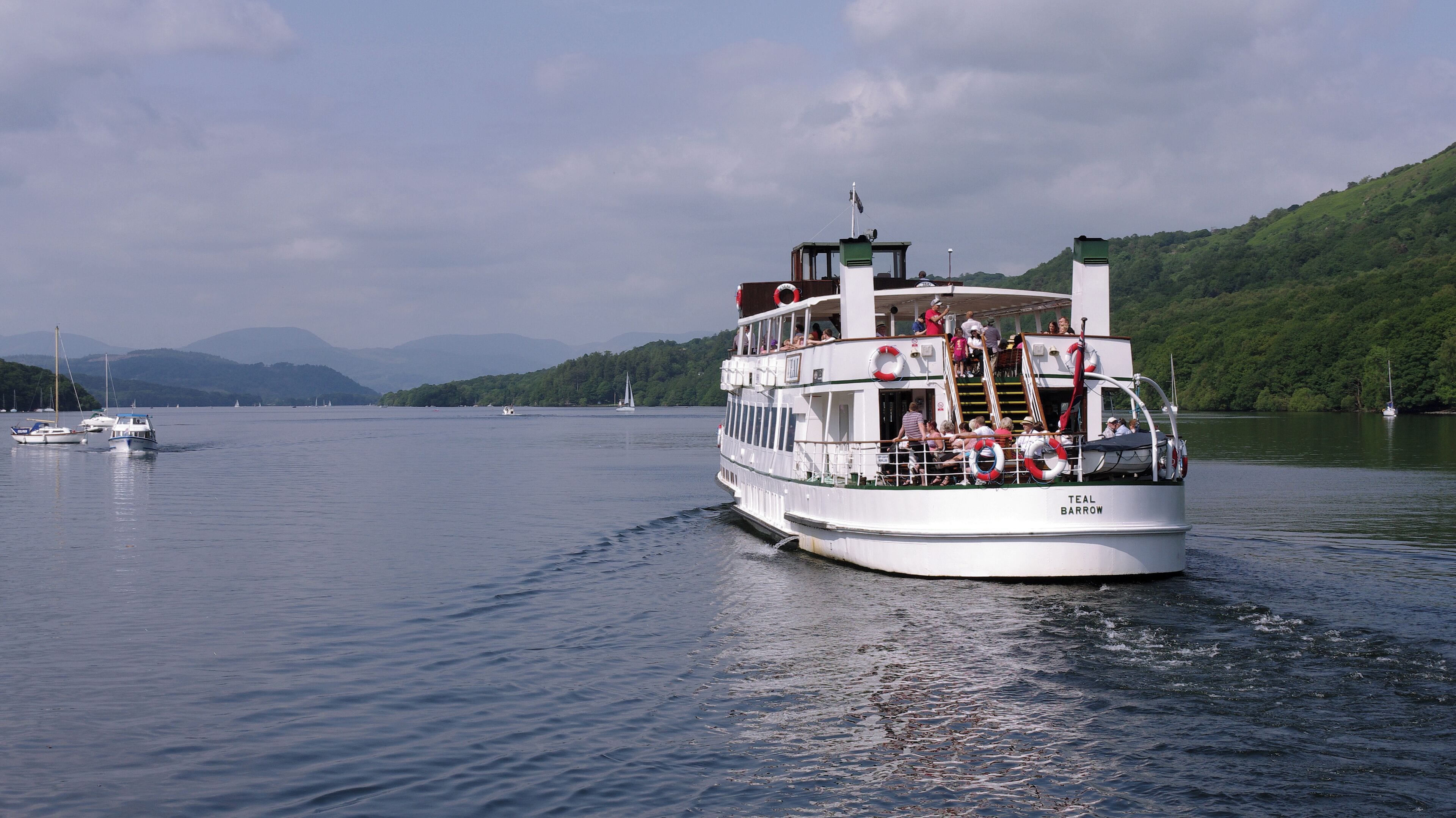 A large white riverboat named "TEAL BARROW" with passengers on deck, moving through a calm river with sailboats in the background and green hills on the opposite bank.