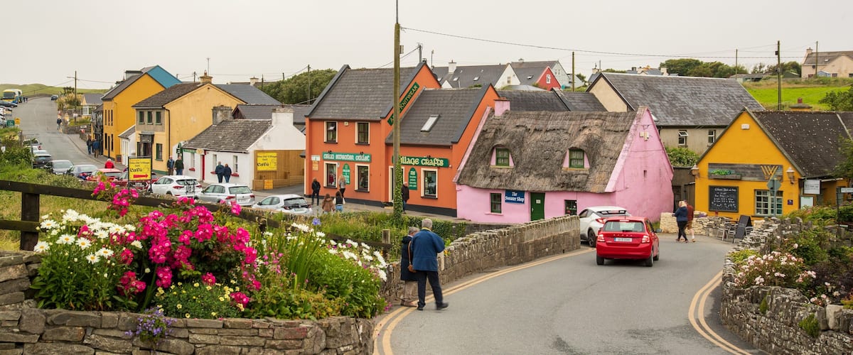 A quaint village street lined with colorful houses, a stone wall with flowers, and a red car driving down the road.