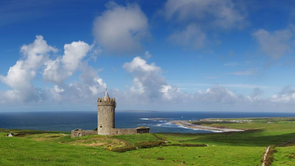 A coastal landscape with a stone tower, green fields, and the ocean.
