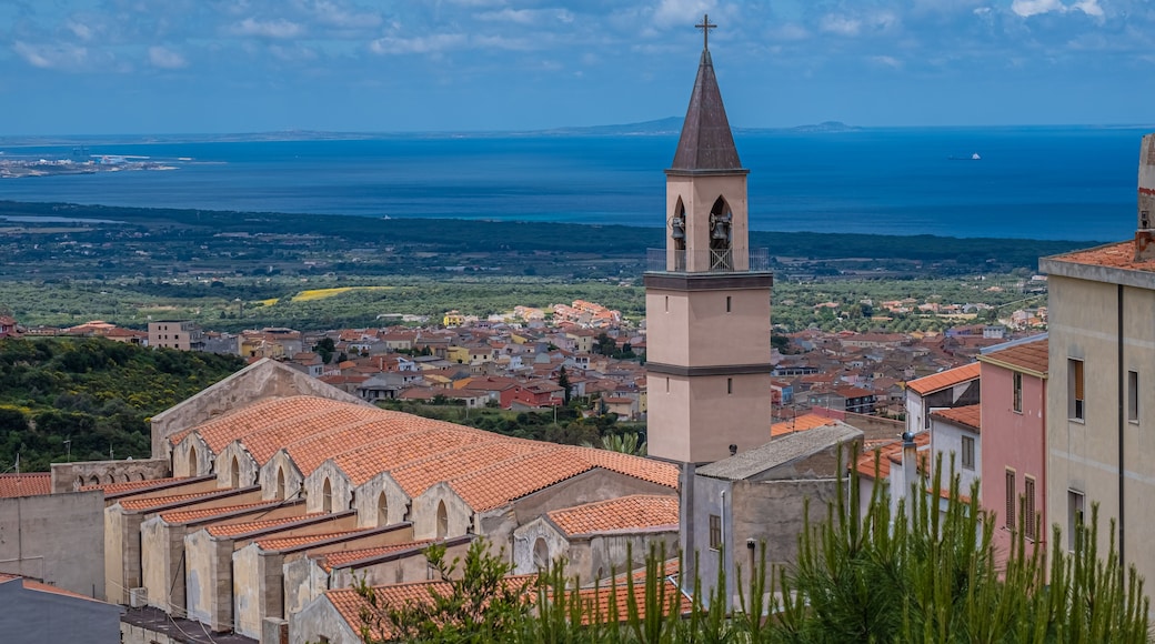 A coastal town with a prominent church tower, terracotta-roofed buildings, and a view of the sea and distant mountains.