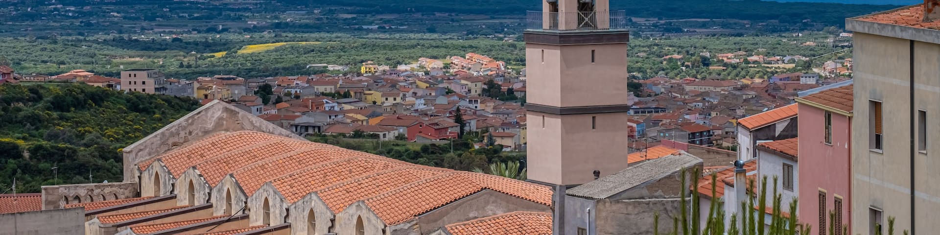 A coastal town with a prominent church tower, terracotta-roofed buildings, and a view of the sea and distant mountains.