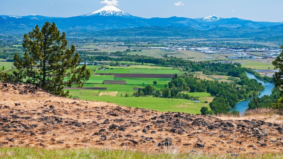 A mountainous landscape with a river, green fields, and a prominent snow-capped peak in the background.