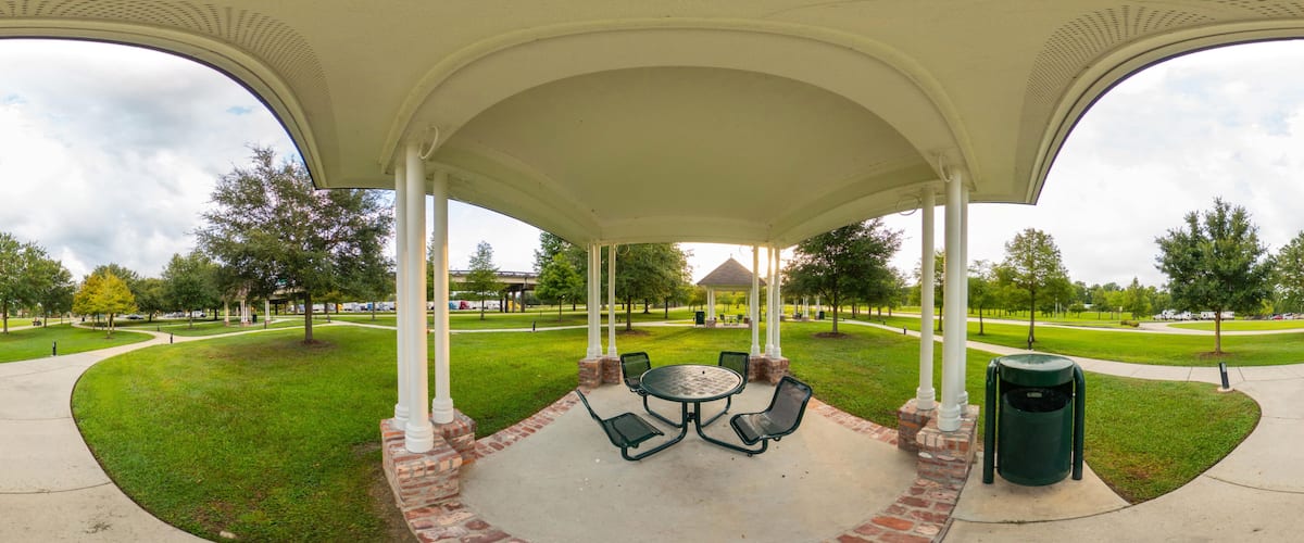 A covered pavilion with a round table and chairs, surrounded by a brick border, set in a park with grass and trees.