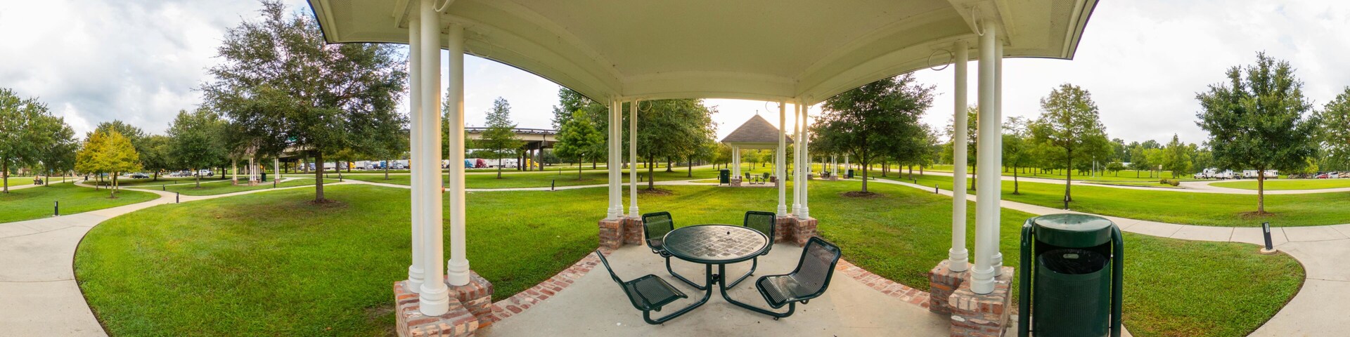 A covered pavilion with a round table and chairs, surrounded by a brick border, set in a park with grass and trees.