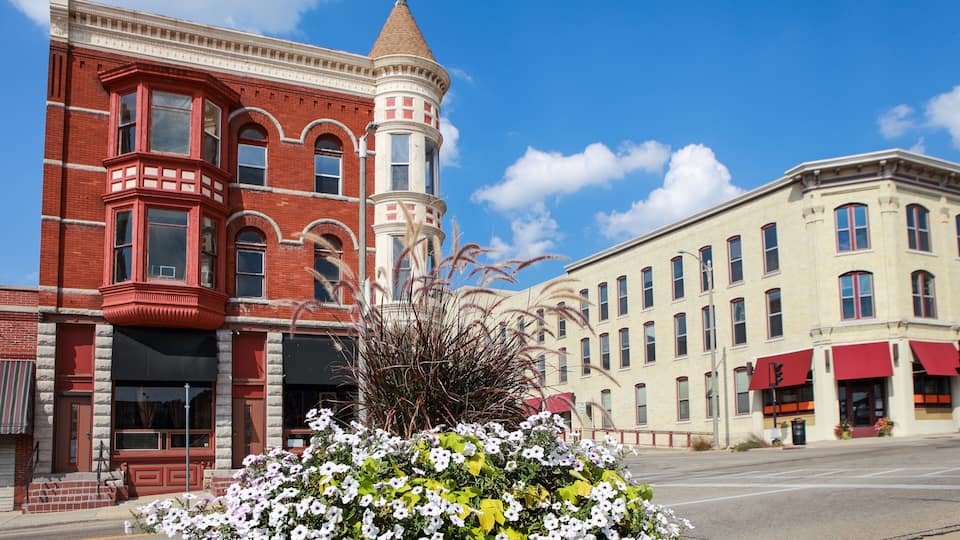 A historic building with a red facade and a corner turret, a street with a flower bed, and another building with a yellow facade and awnings.