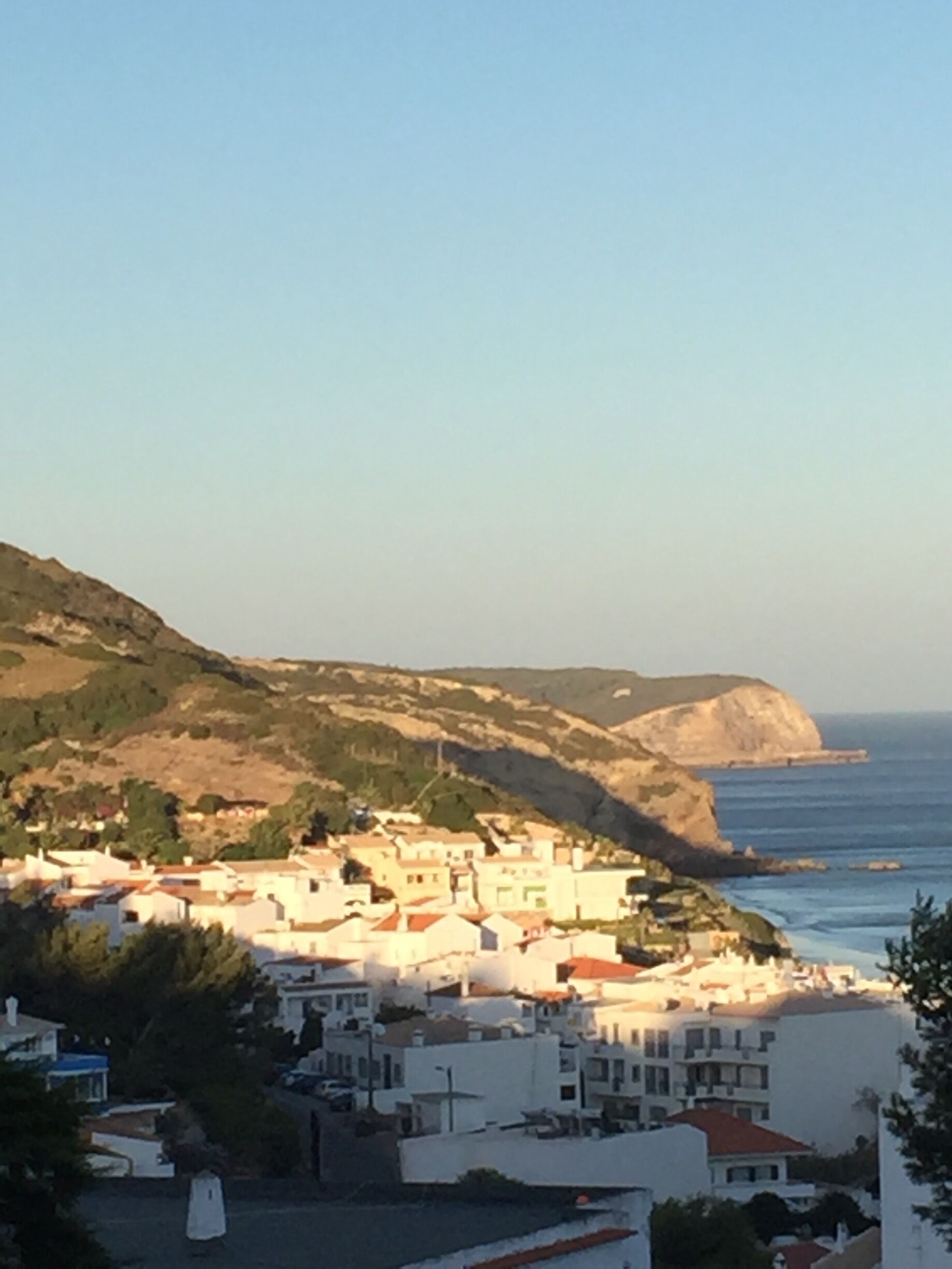 A coastal village with white buildings, a hilly backdrop, and the sea in the distance.