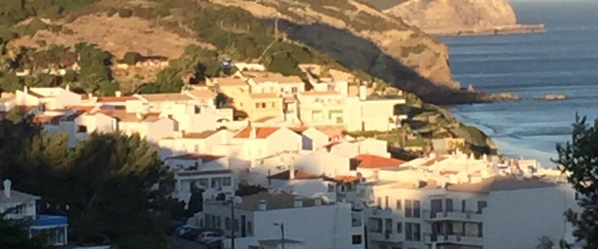 A coastal village with white buildings, a hilly backdrop, and the sea in the distance.