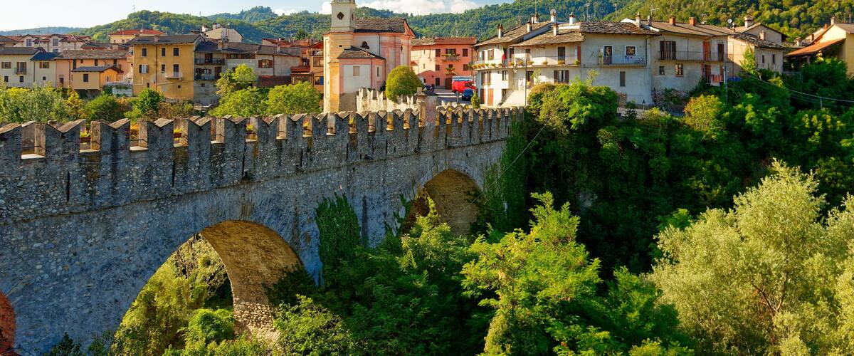 A stone bridge with arches, a tower, and buildings in a valley surrounded by greenery.