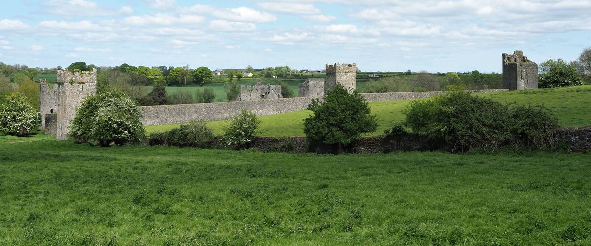 A historic castle ruin with stone walls and towers, surrounded by green fields and trees under a partly cloudy sky.