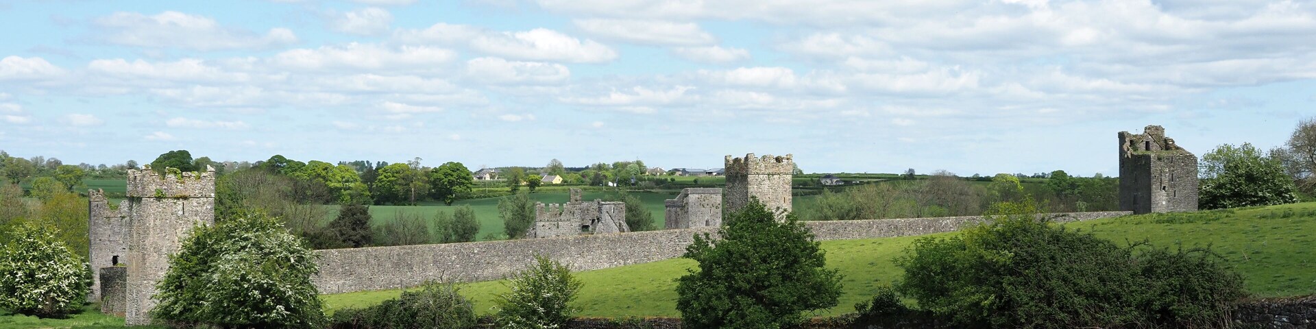 A historic castle ruin with stone walls and towers, surrounded by green fields and trees under a partly cloudy sky.