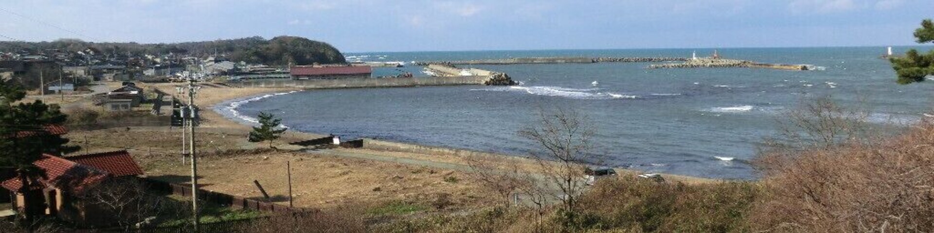 Un paisaje costero con una orilla curva, una playa arenosa y un pequeño muelle que se adentra en el agua.