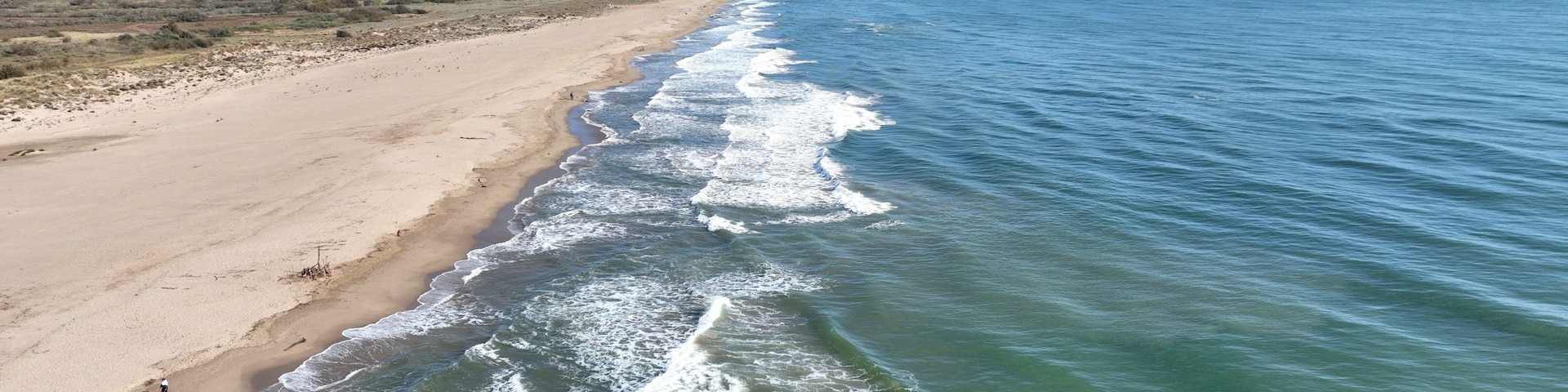 Una playa arenosa con olas rompiendo en la orilla, cielo despejado y una silueta urbana distante.