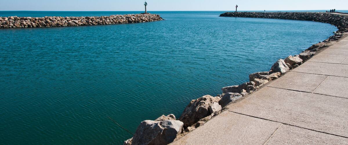 A coastal scene with a stone pier extending into turquoise waters, a rocky breakwater, and a clear blue sky.