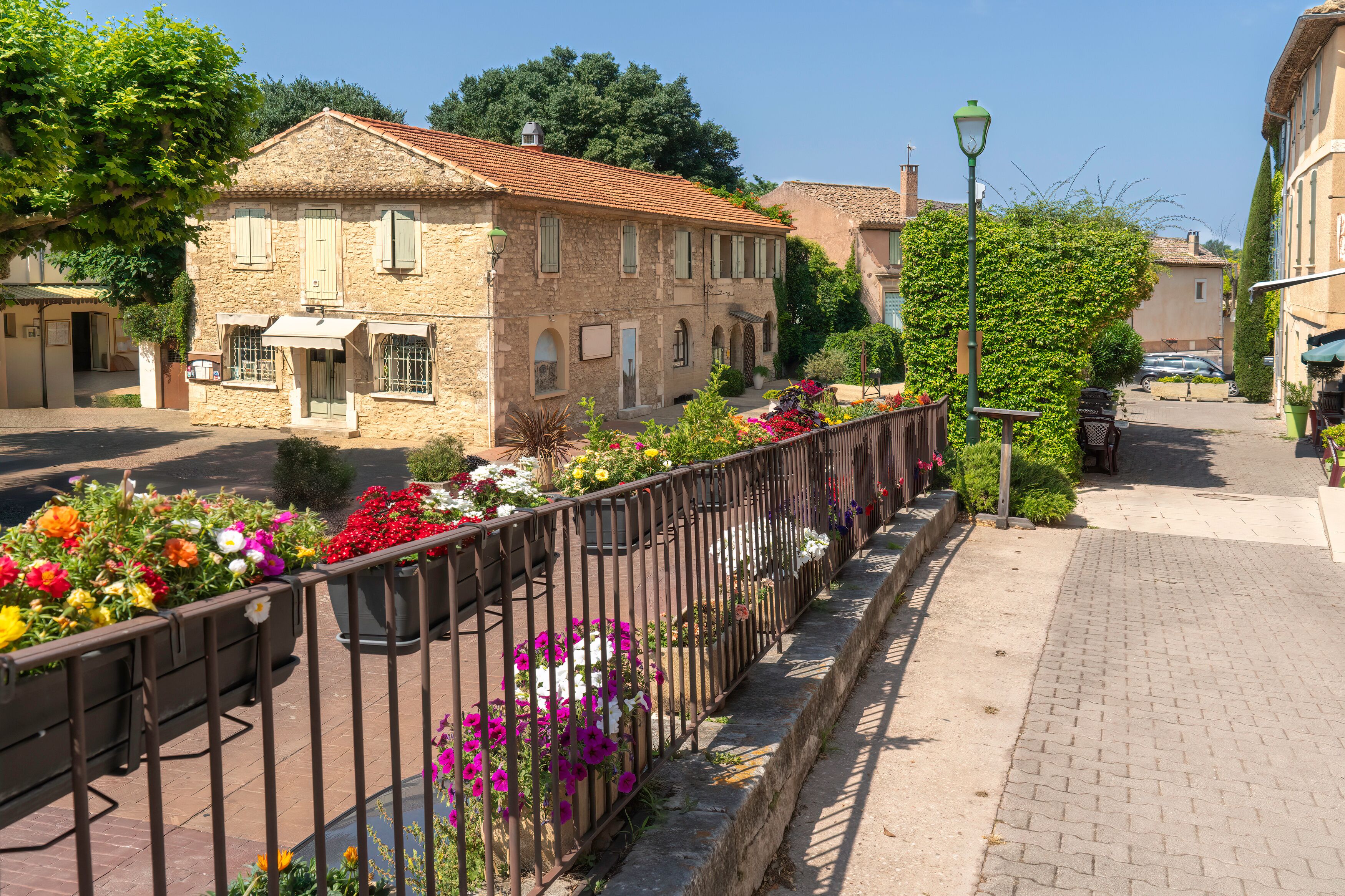 A stone building with a tiled roof, a metal fence with flowers, and a paved sidewalk.