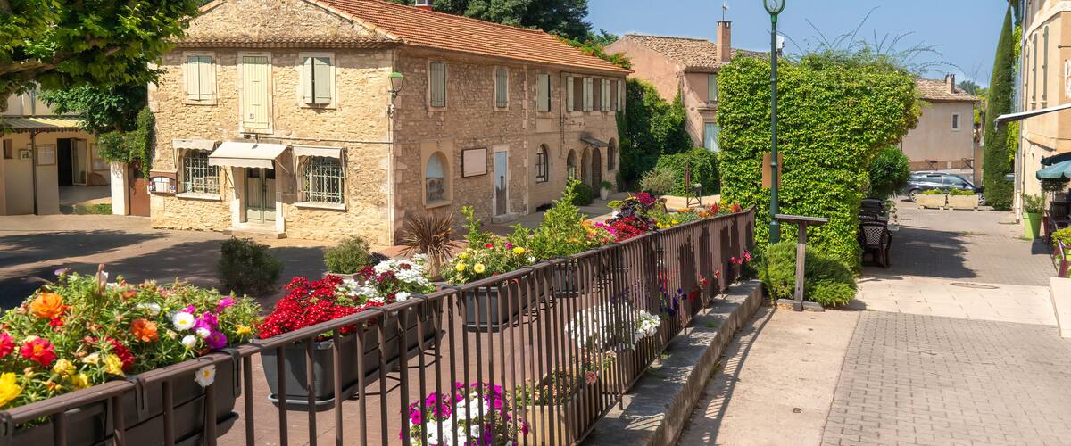 A stone building with a tiled roof, a metal fence with flowers, and a paved sidewalk.