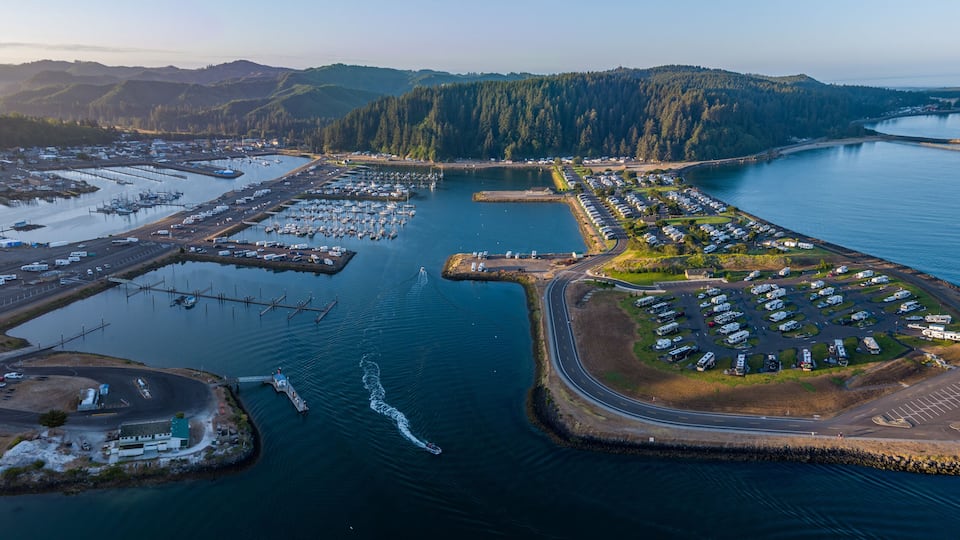 An aerial view of a coastal marina with boats, a parking area, and a forested peninsula.