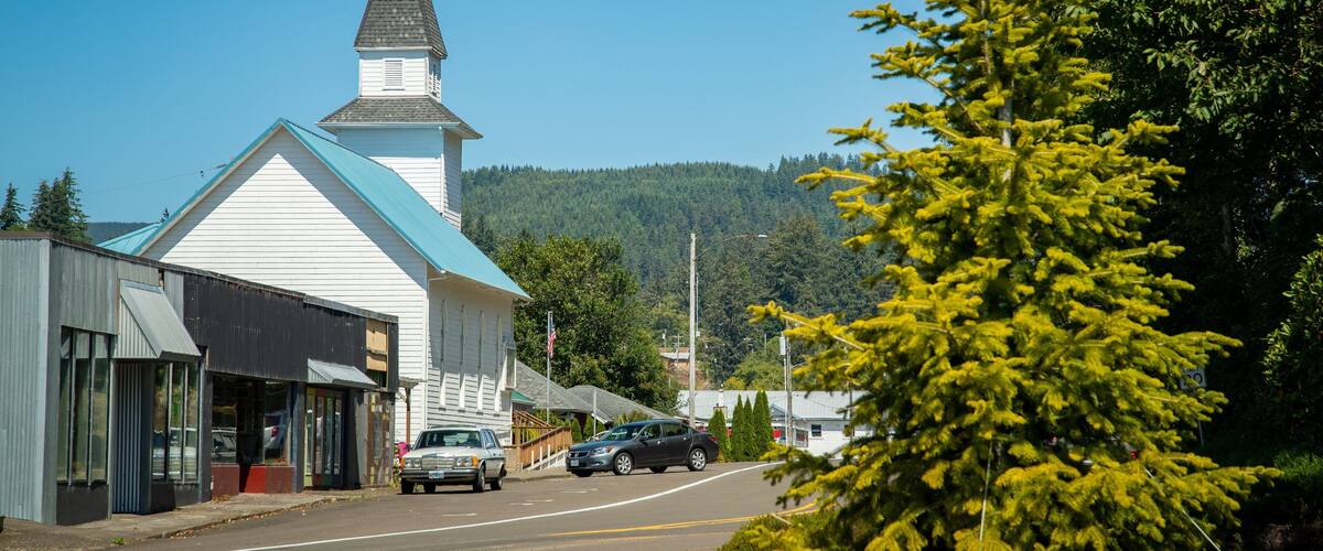 A church with a steeple, a road with cars, and a forested hill in the background.