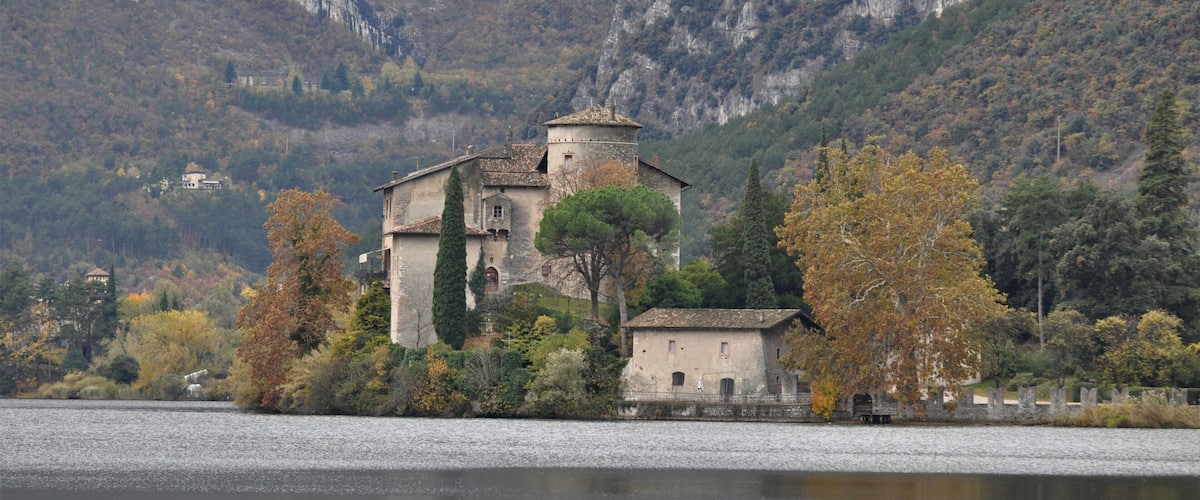A historic building with a tower by a lake, surrounded by autumn trees and a mountainous backdrop.