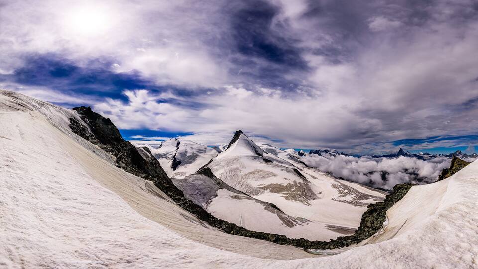A snow-covered mountain range with a valley and rocky peaks under a partly cloudy sky.