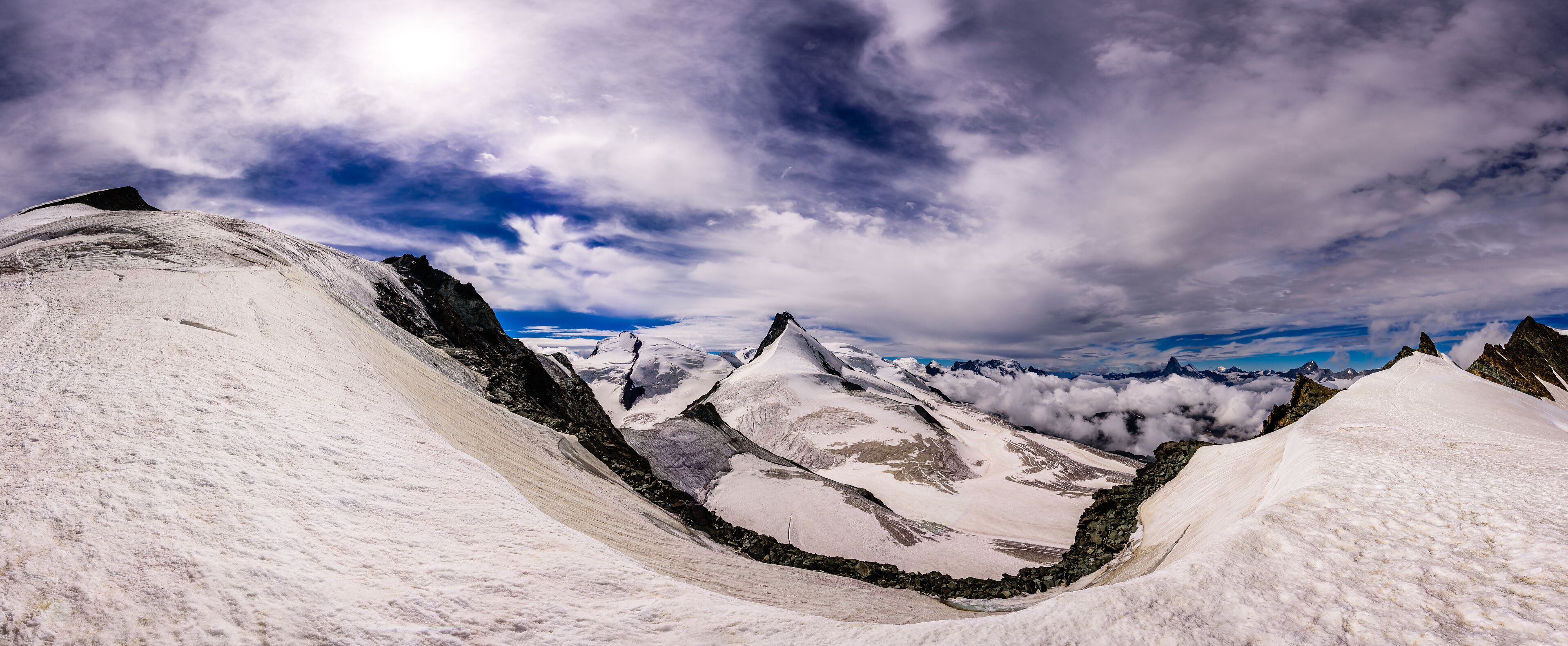A snow-covered mountain range with a valley and rocky peaks under a partly cloudy sky.