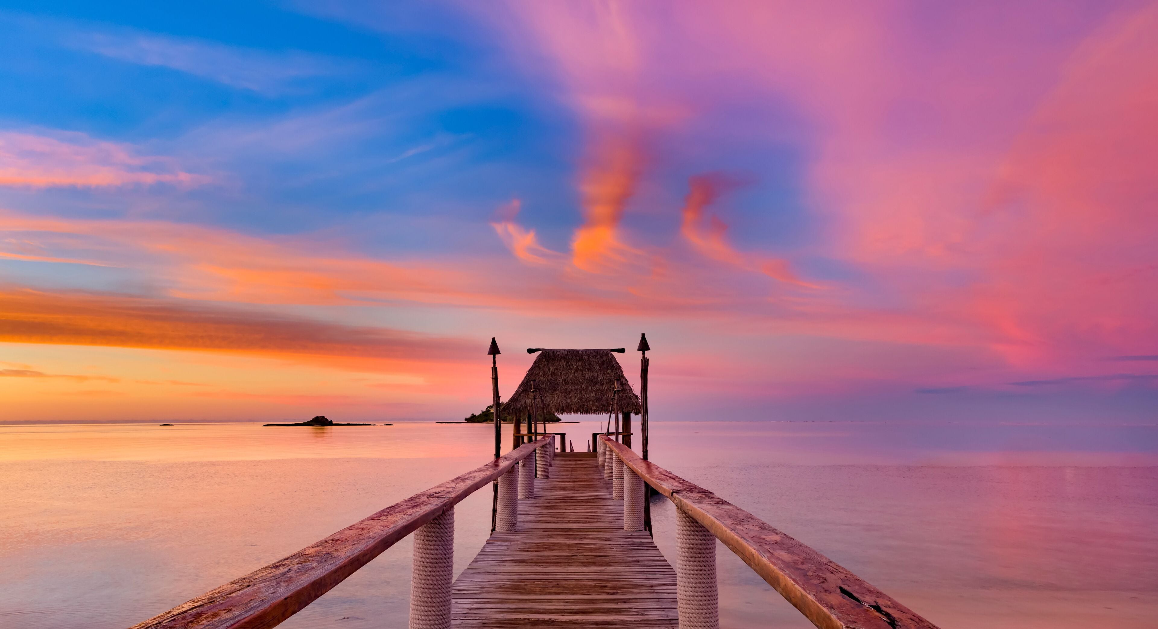 A wooden pier leading to a thatched-roof structure, extending into calm waters with a colorful sunset sky.