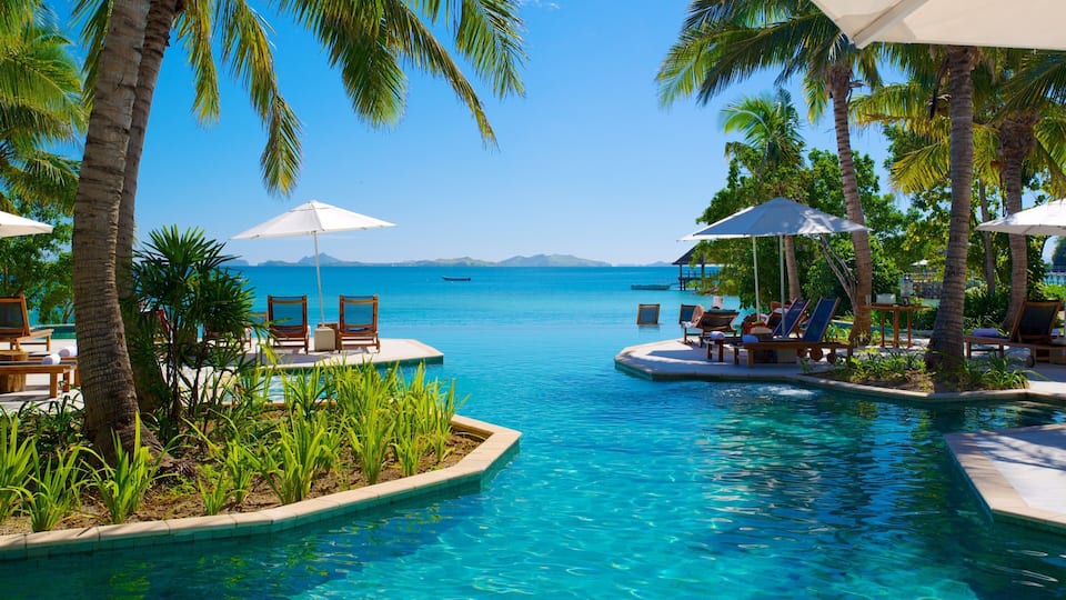 A poolside with lounge chairs and umbrellas, palm trees, and a clear view of the ocean.