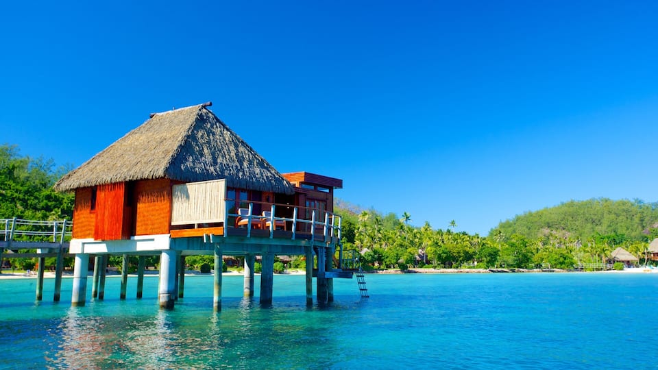 A thatched-roof overwater bungalow with a wooden deck and white chairs, surrounded by clear turquoise water and lush greenery.