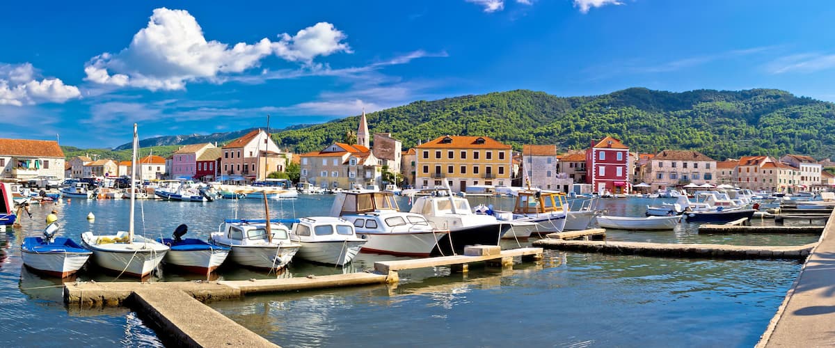 A marina with boats docked, colorful buildings, and a mountainous backdrop.