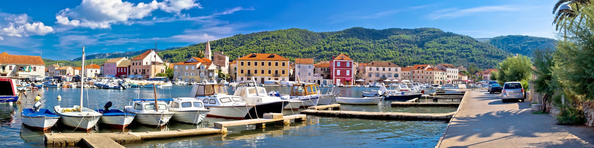 A marina with boats docked, colorful buildings, and a mountainous backdrop.