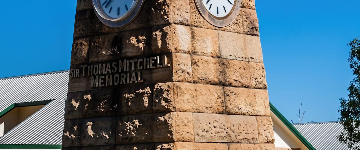 Un campanile in pietra con due orologi, una targa che ricorda 'Sir Thomas Mitchell Memorial' e un caffè sullo sfondo.