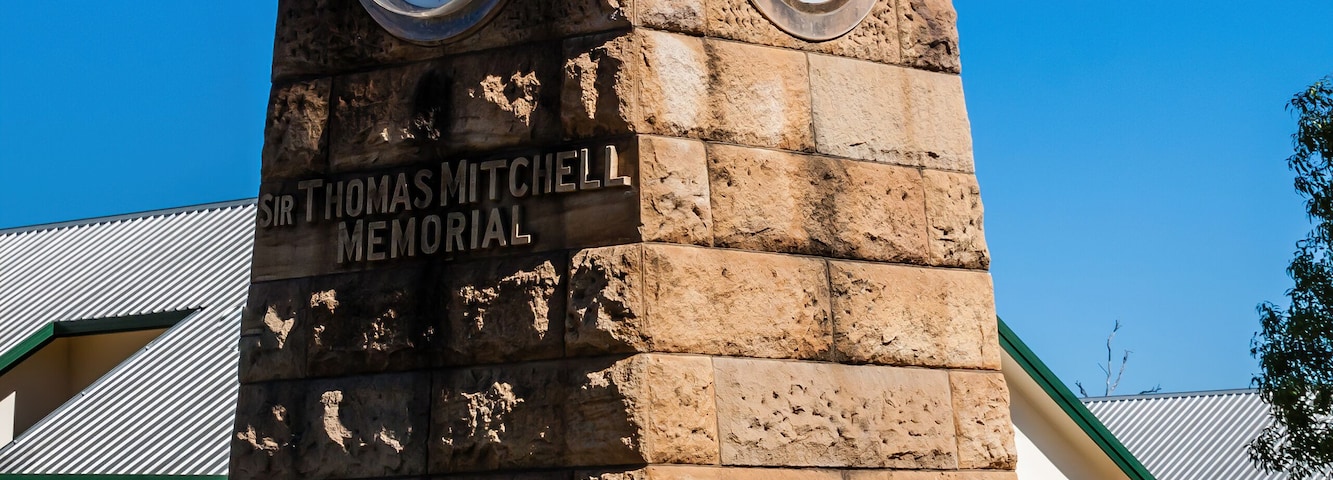 Una torre de reloj de piedra con dos relojes, una placa con la inscripción "Sir Thomas Mitchell Memorial" y una cafetería al fondo.