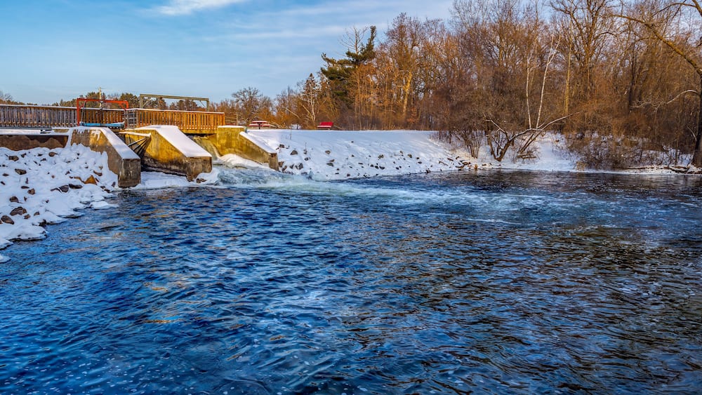 A river flowing under a bridge with snow on the banks and trees in the background.