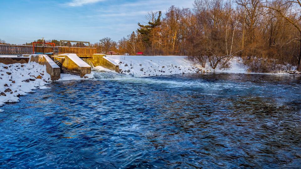 A river flowing under a bridge with snow on the banks and trees in the background.