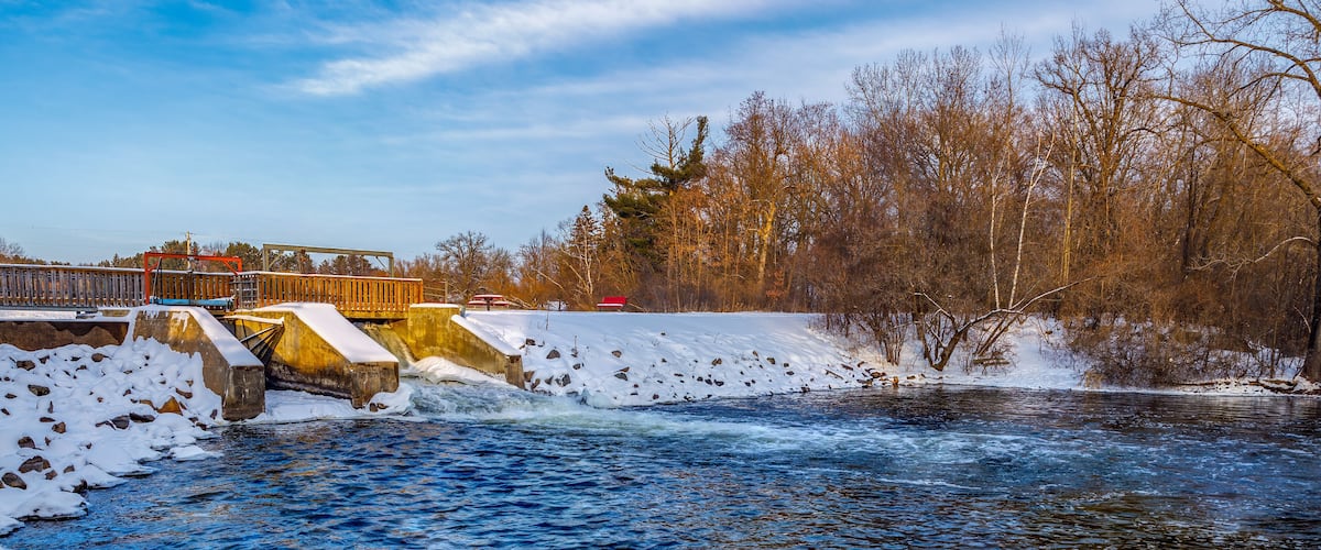 A river flowing under a bridge with snow on the banks and trees in the background.