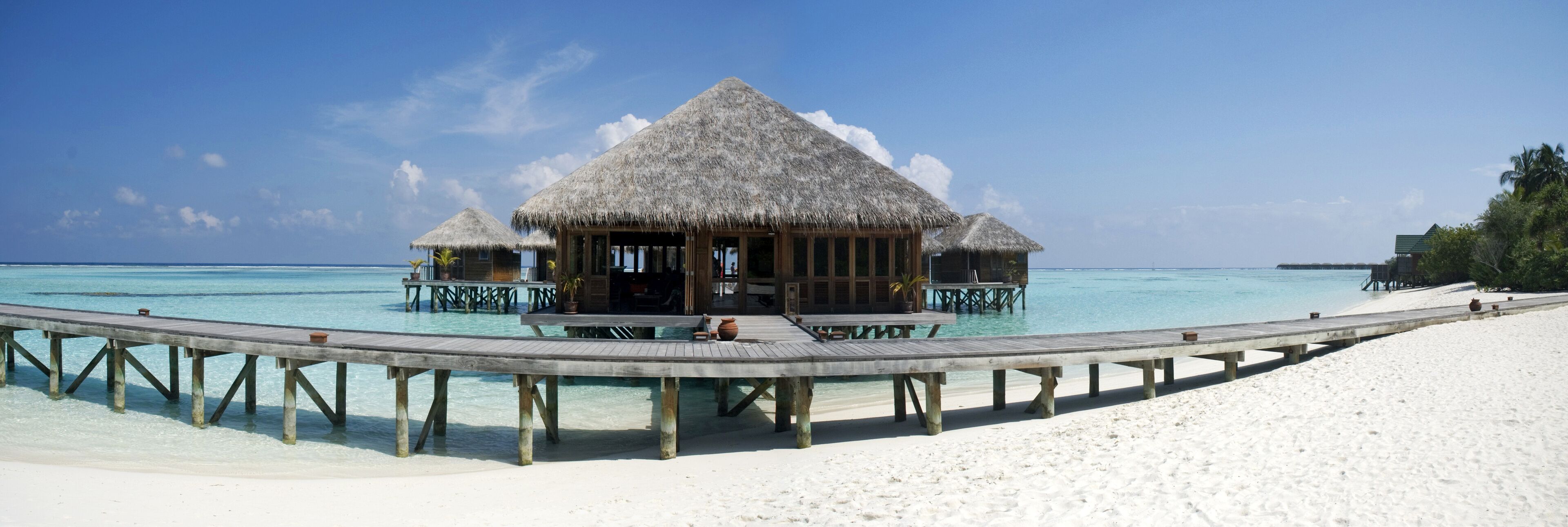 Overwater bungalows with thatched roofs on stilts, a wooden walkway, and a clear blue sea.