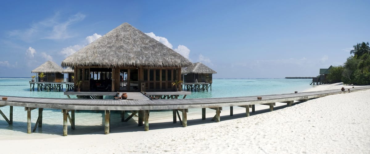Overwater bungalows with thatched roofs on stilts, a wooden walkway, and a clear blue sea.