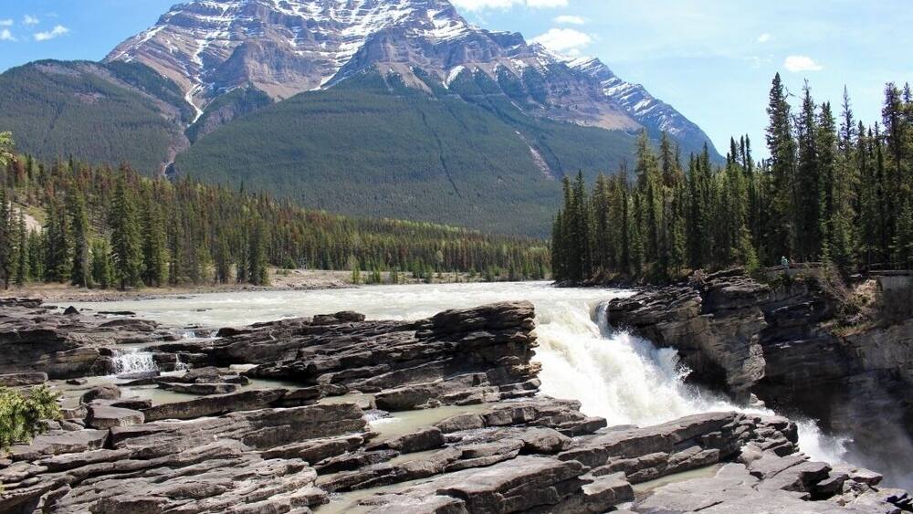 A river flowing over rocky terrain with a waterfall, surrounded by dense forests and mountains in the background.