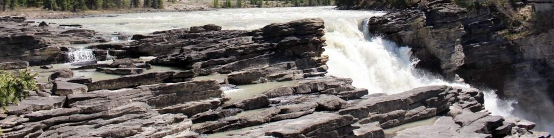 A river flowing over rocky terrain with a waterfall, surrounded by dense forests and mountains in the background.