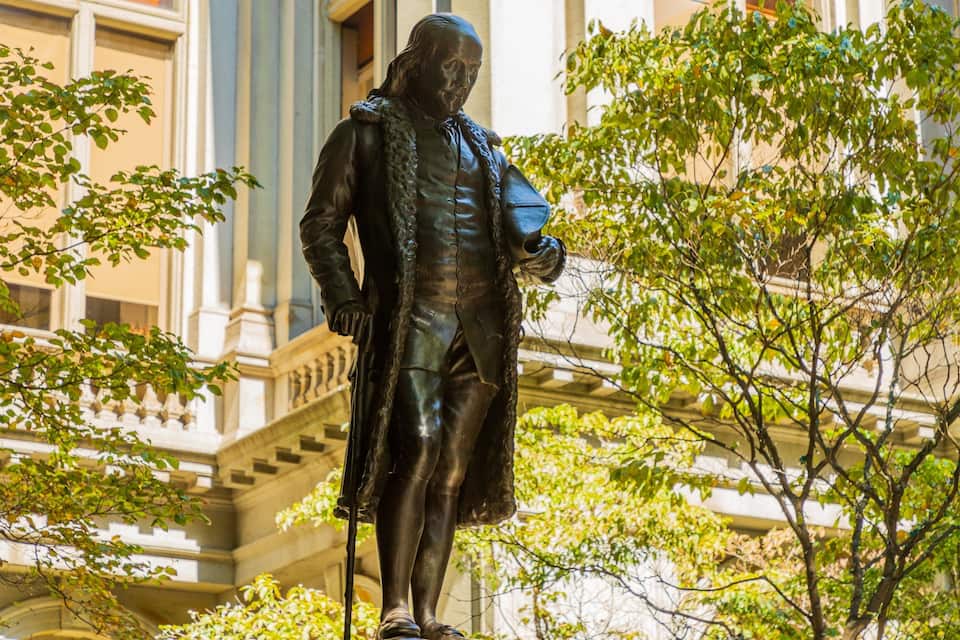 A statue of a historical figure in period clothing, surrounded by trees and classical architecture.