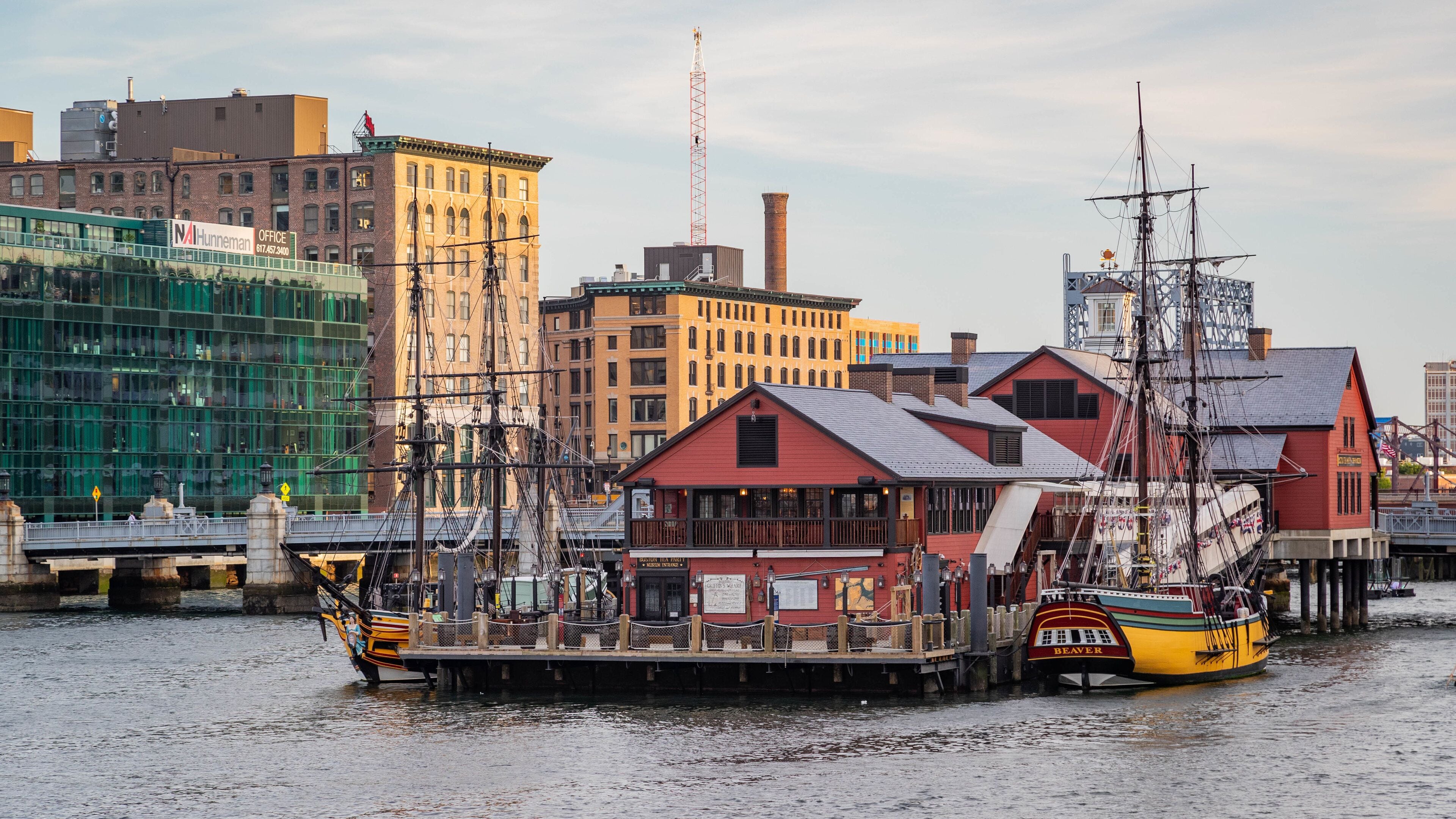Ein historischer Uferbereich mit einem hohen Segelschiff, das an einem Steg liegt, umgeben von Industriegebäuden und einer Brücke.