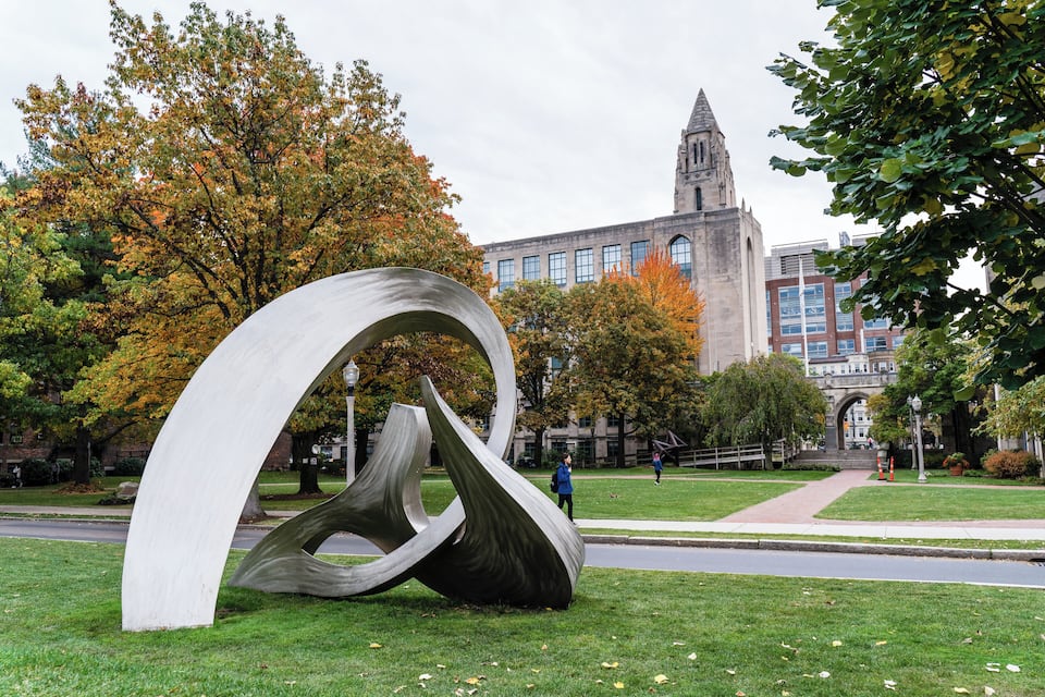 A modern sculpture in a park with autumn trees and a historic building in the background.