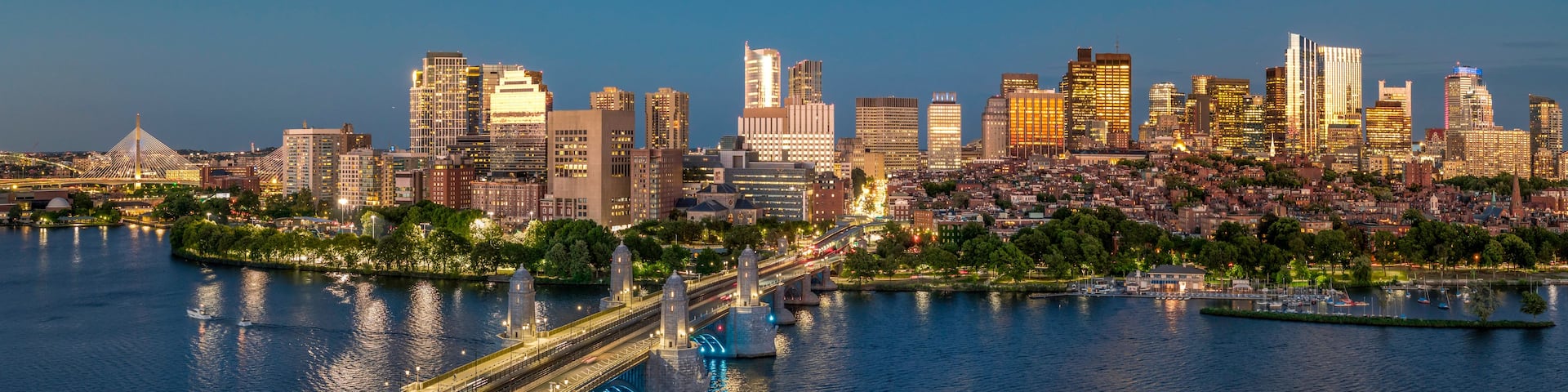 A city skyline at dusk with illuminated buildings, a river, and a bridge.